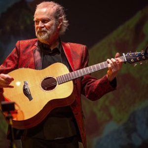 Beppe Gambetta performing live acoustic guitar on stage in a red jacket at Locktown Stone Church concert.